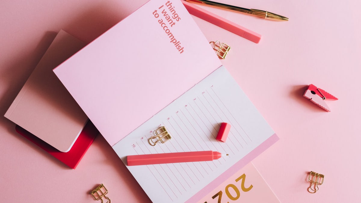 Organized workspace with pink planner, pen, and clips on pastel background.